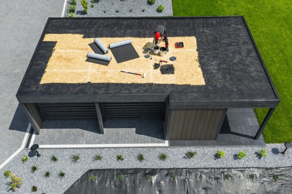 A team is working on the roof of a contemporary building, applying EPDM roofing materials while surrounded by a well-kept yard and clear skies.